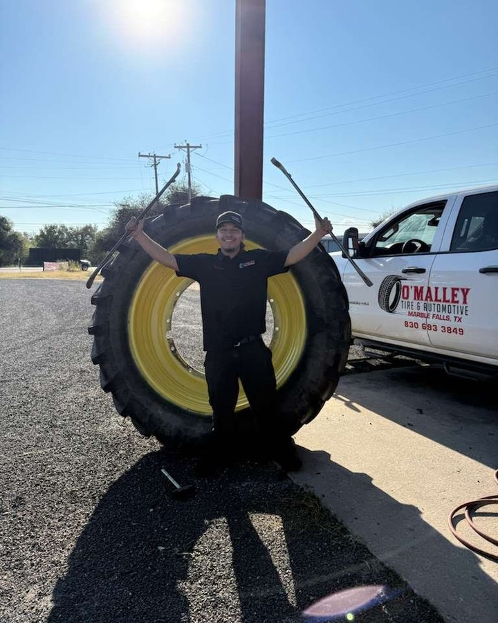 Male mechanic posing with a giant tractor tire, holding tire irons in both hands beside a service truck under a sunny sky.