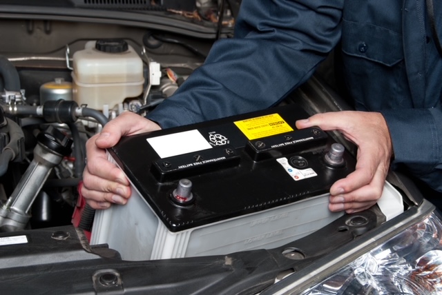 Technician in blue coveralls holding a car battery in a vehicle's engine bay during battery replacement.