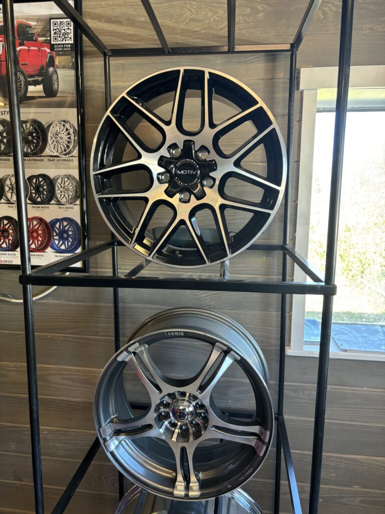 Large black-and-silver alloy wheel on a display shelf in a tire shop, center cap reads MOTIV rim brand.
