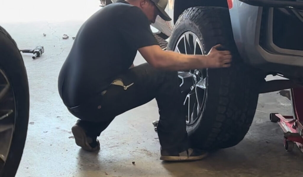 Man crouched beside a raised SUV, inspecting a large tire during a tire change in a garage.
