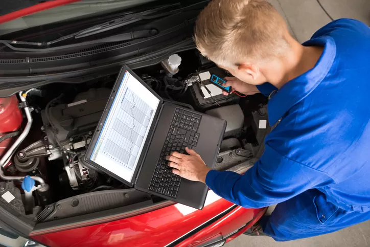 Auto technician in blue coveralls uses a laptop to diagnose a red car under the hood, with diagnostic cables nearby.