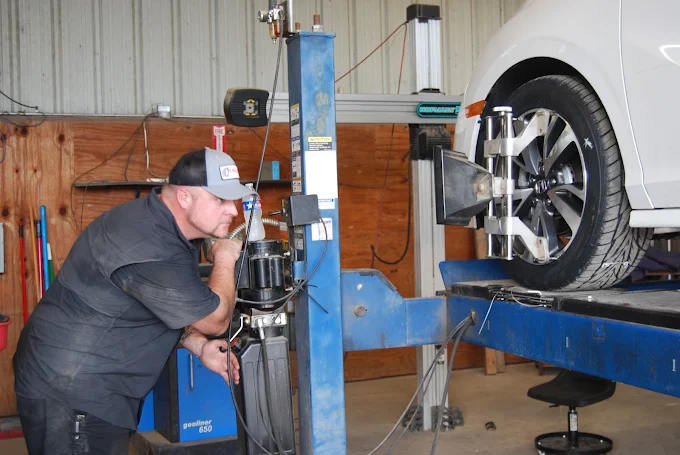 Auto mechanic wearing a cap inspects a car elevated on a blue hydraulic lift inside an auto repair shop.