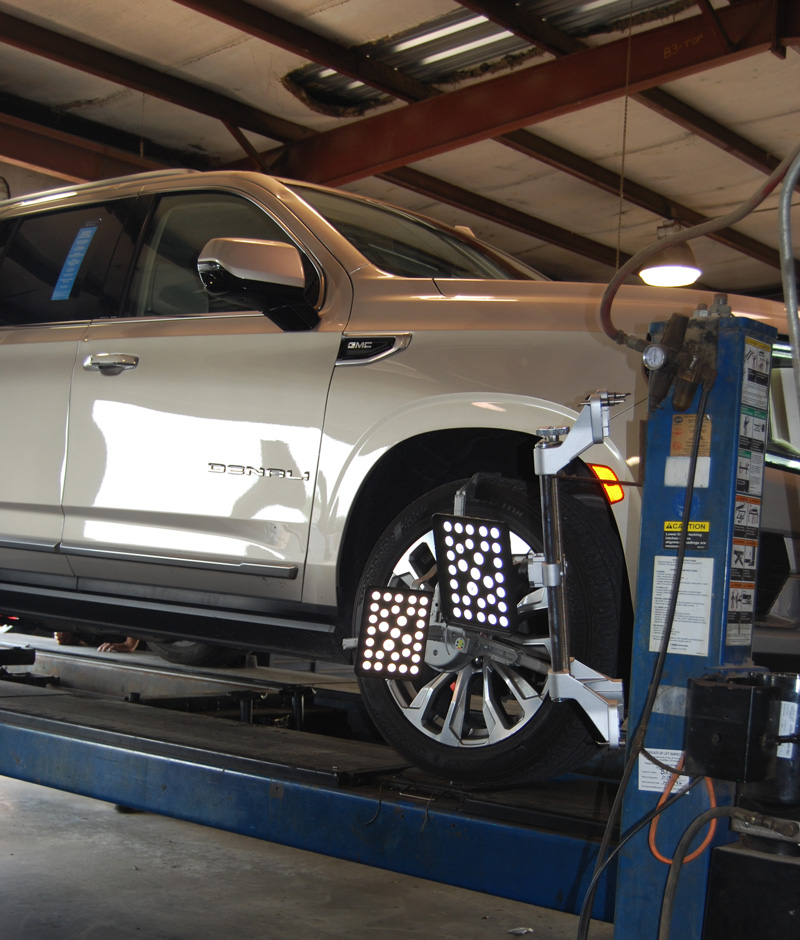 White GMC Denali SUV on a car lift inside an auto repair shop, with diagnostic lighting panels positioned near the front wheel.