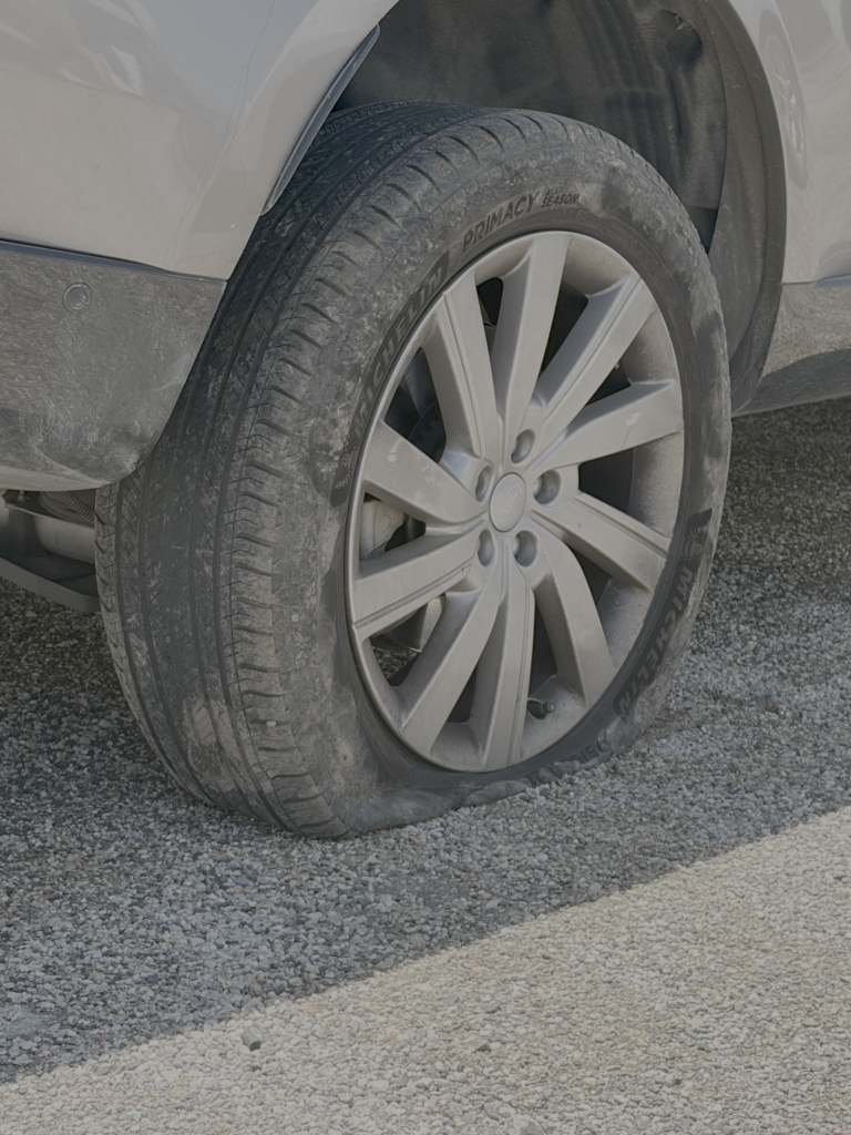 Close-up of a dusty car tire and alloy wheel, resting on a gravel surface beside a light-colored vehicle.