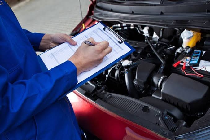 Mechanic in blue overalls writing on a clipboard while inspecting an open car hood