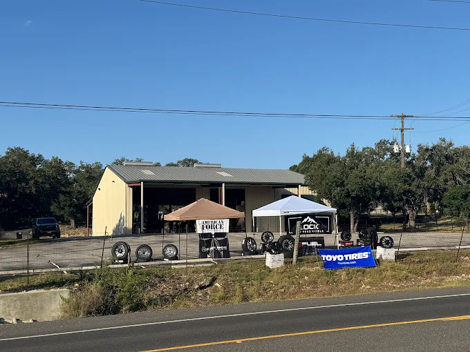 Tire display along a fence in front of a beige auto shop, with two canopies (brown and white) and stacked wheels lined up outside on a sunny day