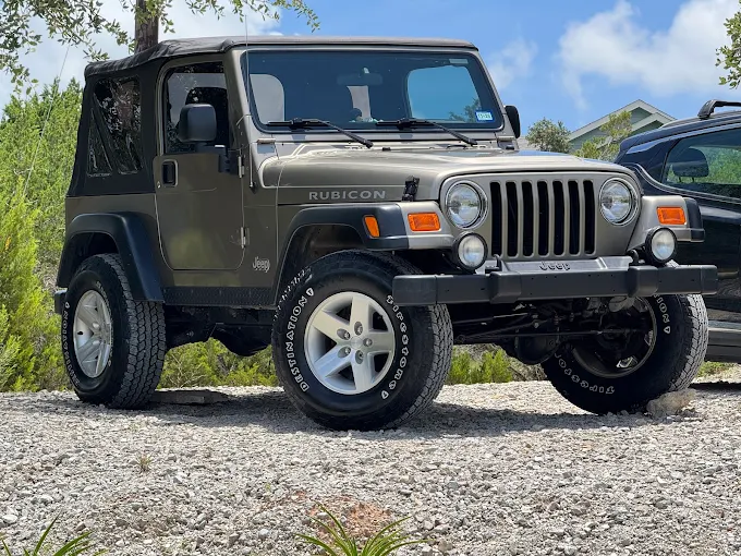 Olive-green Jeep Wrangler Rubicon with black soft top, front three-quarter view parked on gravel.
