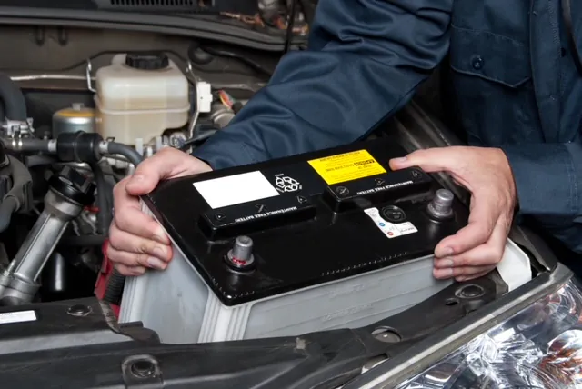 Technician in blue coveralls holding a car battery in a vehicle's engine bay during battery replacement.
