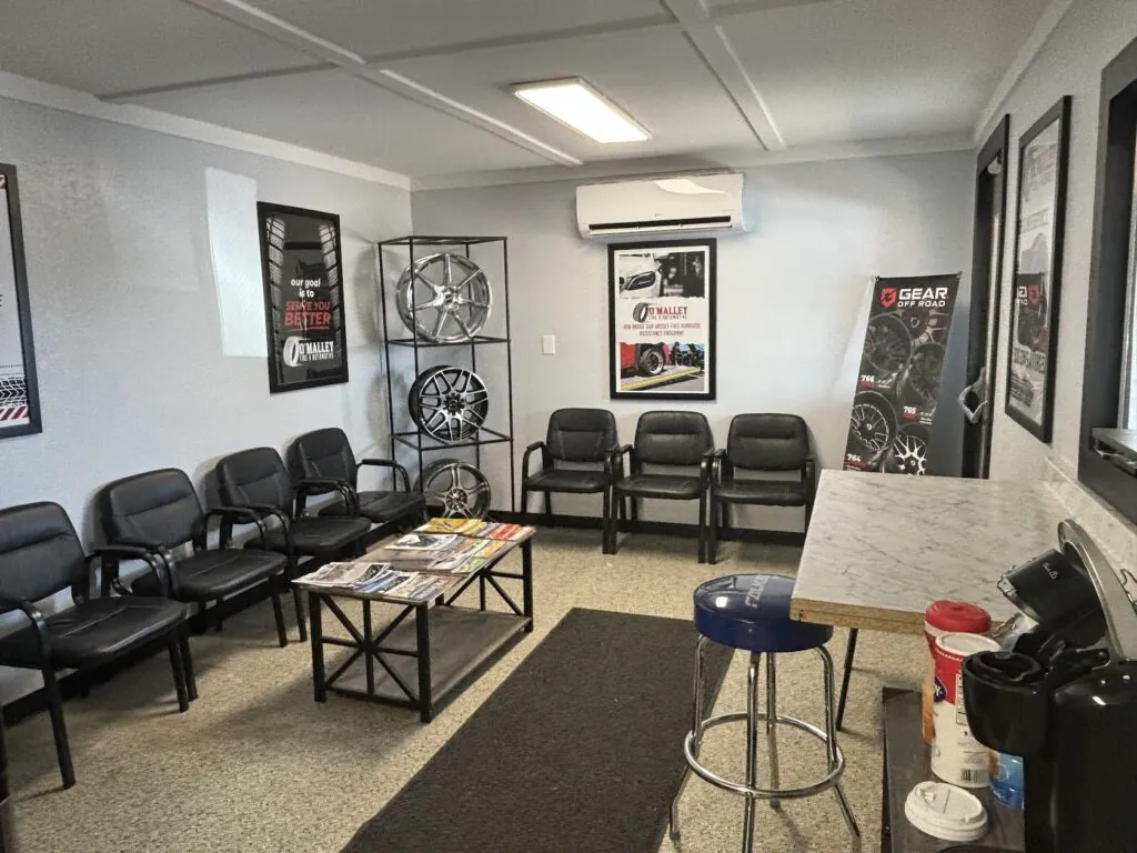 Car showroom waiting area with black chairs, a display rack of alloy wheels, framed posters, and a marble-front counter with bar stools.
