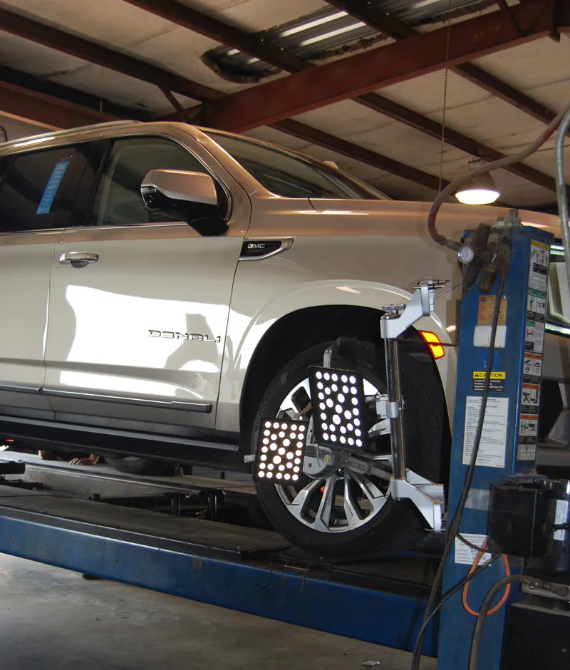 White GMC Denali SUV on a car lift inside an auto repair shop, with diagnostic lighting panels positioned near the front wheel.