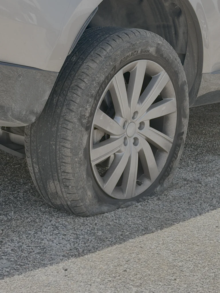 Close-up of a dusty car tire and alloy wheel, resting on a gravel surface beside a light-colored vehicle.